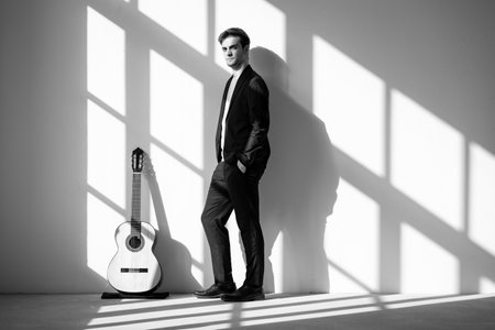 Portrait of a young boy standing next to his guitar, his hands in his pockets. He is wearing a black suit. Interior of a renovated former factory.の写真素材