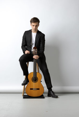 Young boy sitting on the stool in a black suit, holds his wooden classical guitar with one hand. White backgroundの写真素材