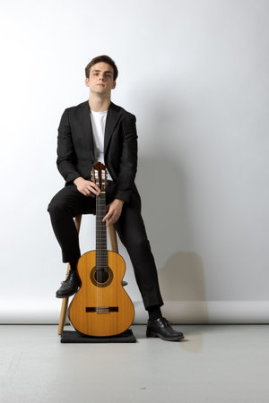 Young boy sitting on the stool in a black suit, holds his wooden classical guitar with one hand. White backgroundの写真素材