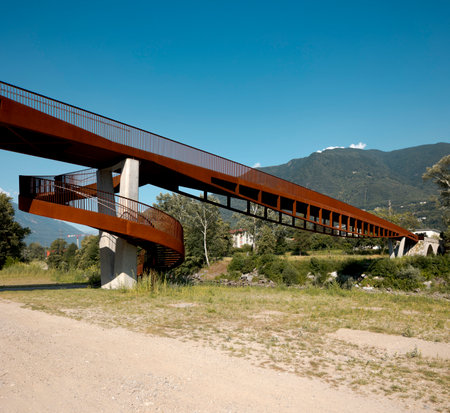 Modern rusted pedestrian and bicycle bridge with flowing Ticino river below. Mountain panorama of the Swiss Alps on a sunny summer day. No one insideの写真素材
