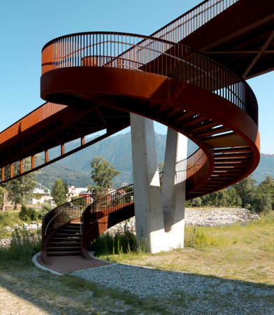 Modern rusted pedestrian and bicycle bridge with flowing Ticino river below. Detail of the spiral staircase and mountain panorama of the Swiss Alps in the background. No one insideの写真素材
