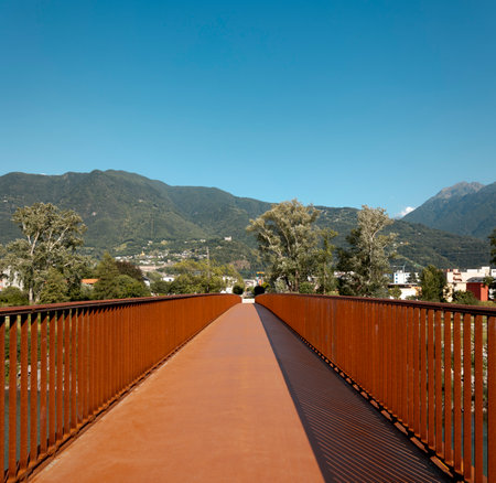Front view of the pedestrian and bicycle path of a modern rusty bridge, with the Swiss Alps in Ticino in the background. Strong contrast between light and shadow, nice panorama. No one insideの写真素材