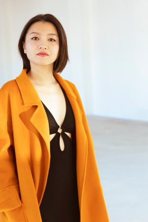 Portrait of a young Chinese woman wearing a nice orange coat and a black skintight dress. Photographed inside a white factory.の写真素材