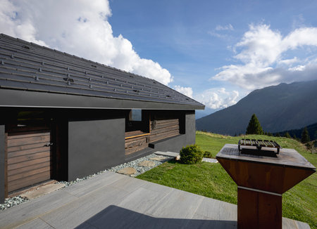 A very unusual Swiss mountain chalet because it is black and made of wood. Surrounded by a beautiful green summer meadow. The sky is blue and it looks like a quiet and very peaceful place. A very modern grill for cooking meat outside.の写真素材
