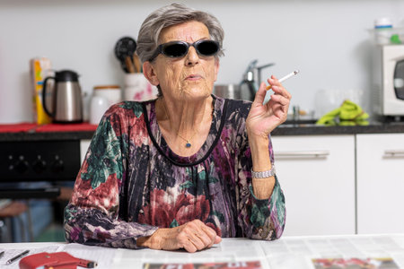 Portrait of old smoker sitting in her modern white kitchen with dark sunglasses. A woman from...