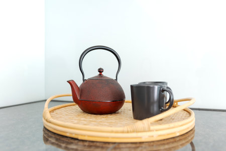 Detail of a red Chinese teapot and two black cups resting on a wicker tray, in a bright kitchen. No one insideの写真素材