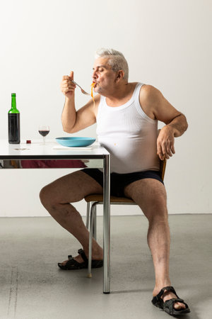 Portrait of a man in underwear and a white undershirt soiled with sauce, sitting at a table while eating a plate of spaghetti with tomato sauce. Isolated scene of domestic lifeの写真素材