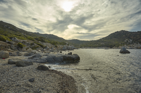 Seascape al tramonto contenente una magnifica spiaggia mediterranea con i promontori rocciosi e le montagne sotto un cielo nuvoloso con spiragli di sole.の写真素材