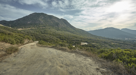 Narrow mountain dirt road with landscape behind rich vegetation under a cloudy sky.の写真素材