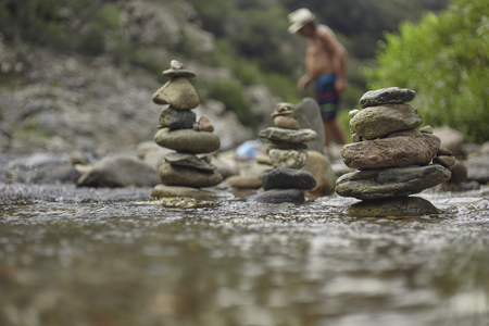 Stacks of zen rocks in a mountain sink with a disfigured man in the background that crosses the stream itself.の写真素材