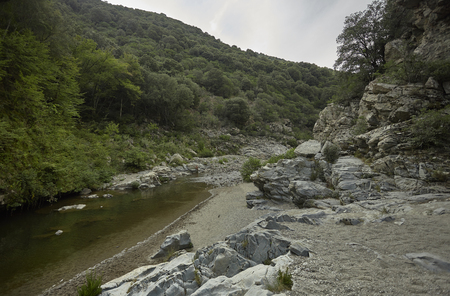Panorama of a stream in the middle of the mountains bordered by a rocky wall and surrounded by green trees that cover the mountains of central Sardinia.の写真素材