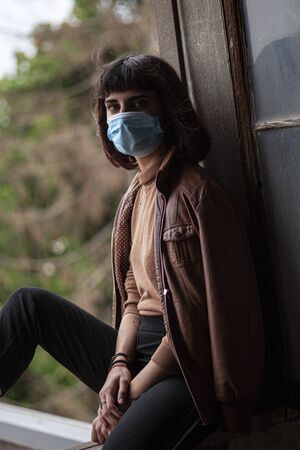 Girl with medical mask at window in her home during covid quarantine periodの写真素材