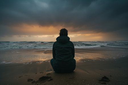 A moody silhouette of a young person sitting alone on a deserted beach, gazing at the waves crashing against the shore.の素材