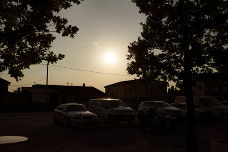 Milan, Italy 12 June 2024: A serene sunset over a suburban street, with the sun low in the sky casting a warm, golden light. The scene includes a few parked cars and motorcycles, with silhouettes of housesのeditorial素材
