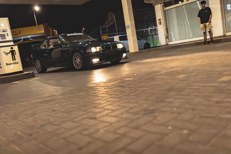 BMW E36 with custom wheels and headlights on, parked at a gas station, showcasing its sporty design