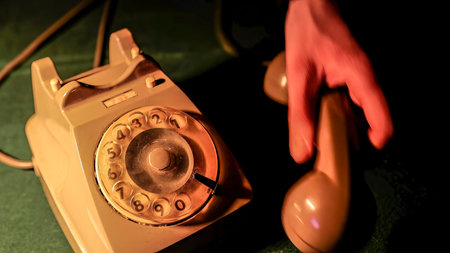 Close-up of hand picking up the receiver of an old rotary phone, illuminated by dramatic lighting,...