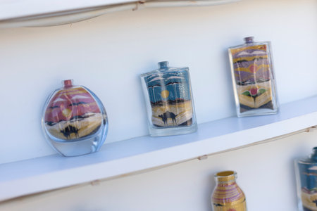 Glass bottles filled with colorful sand art depicting desert scenes are displayed on white shelves, showcasing a traditional Egyptian craft in Marsa Alamの写真素材