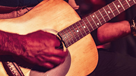 Close-up of a musician's hands playing an acoustic guitar on a stage, bathed in dramatic red...