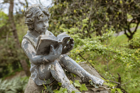 Weathered statue of young child reading book in serene garden setting, enjoying literature amidst natureの写真素材