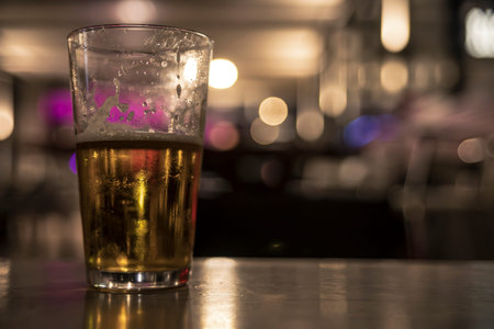 Refreshing cold glass of beer sits on a bar with a blurred background suggesting a lively atmosphere