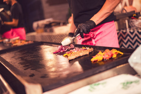 Cook preparing street food with sausage, onions, and peppers using tongs on a hot plate at a food stallの写真素材