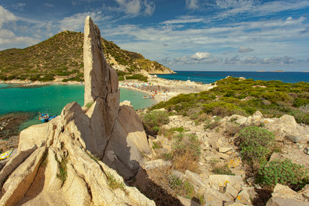 Tourists enjoying the turquoise water and white sand beach of Punta Molentis in Villasimius, Sardinia, with granite rocks and vegetation in the foregroundの写真素材