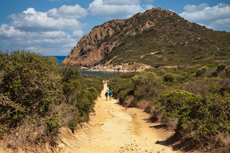 Tourists are walking on a dirt path surrounded by vegetation, leading to the beautiful cala sa figu beach with a rocky promontory in the background, in sardinia, italyの写真素材