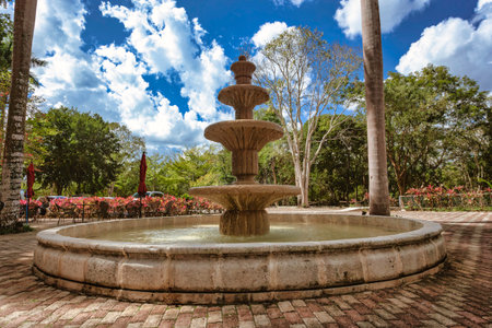 Ornate stone fountain spraying water under a blue sky with clouds, surrounded by lush greenery and palm treesの写真素材
