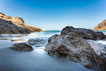 Gentle waves washing the dark sand of cala sa figu beach, with imposing rocks and cliffs creating a picturesque scenery in sardinia, italyの写真素材