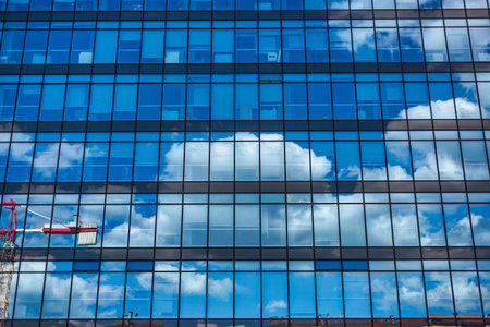 Glass facade of a modern office building reflecting a construction crane and white clouds against a...