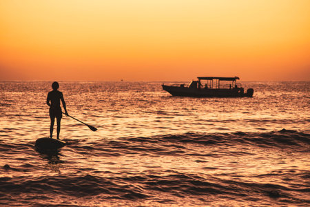 Silhouette of tourist paddle boarding in ocean at sunset with boat in background in dominicus,...
