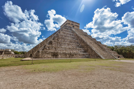Kukulkan temple, an ancient Mayan pyramid, stands tall against a vibrant blue sky dotted with fluffy...