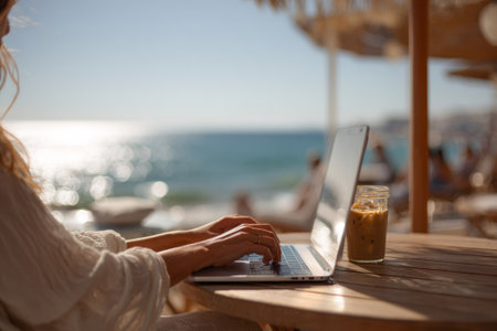 Businesswoman using laptop and enjoying iced coffee at a beachside cafe, embracing the digital nomad lifestyleの素材