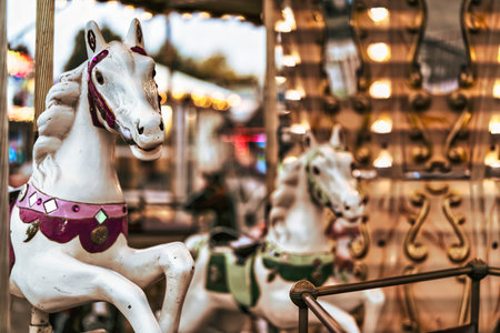 Ornate white carousel horse standing ready for riders with a blurred background displaying the warm glowing lights and golden details of a vintage amusement parkの写真素材