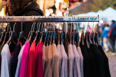 Sweaters of various colors hanging on a clothing rack at an outdoor market stall, with a shopper browsing in the blurred background, creating a scene of retail activity and fashion choicesの写真素材