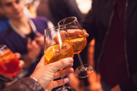 Hands clinking glasses of refreshing spritz with orange slices during a vibrant evening party,...