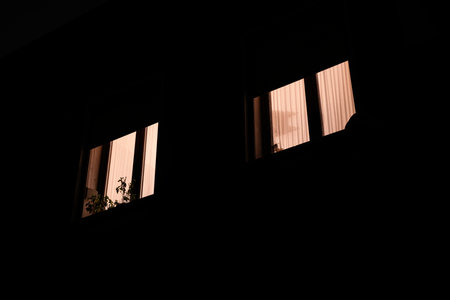 Building facade at night with two warm glowing windows, minimal urban scene evoking mystery, privacy and quiet solitude behind illuminated silhouettes and shadowed wallsの写真素材