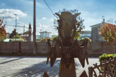 Grotesque stone sculpture featuring an abstract, horned face and weathered texture, standing in an outdoor setting with a sunny sky and blurred urban backgroundの写真素材