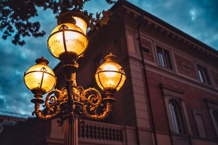 Ornate vintage lamppost standing on an urban street, illuminating the pathway with warm light against a dark blue sky at dusk, a historic building visible in the backgroundの写真素材