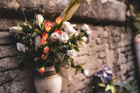 Artificial rose and tulip bouquet in a decorative vase against a weathered stone wall, evoking faded...