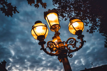 Ornate vintage lamppost stands against a moody overcast sky, casting a warm golden glow, providing...
