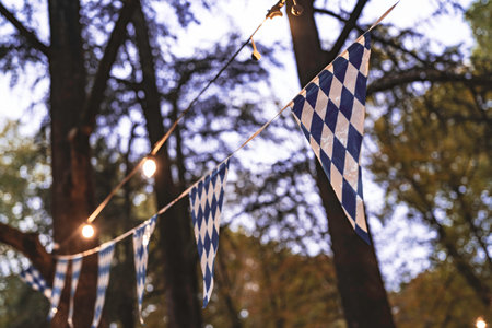 Bavarian blue-and-white diamond bunting and warm string lights drape across a blurred forest, evoking outdoor oktoberfest cheer, autumn warmth, and festive evening atmosphereの写真素材