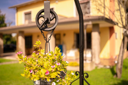 Rustic iron well pulley with a hanging bucket planter full of green plants and small pink flowers, decorating a sunny residential garden with a blurred yellow house in the backgroundの写真素材