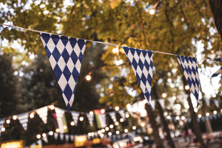 Bavarian diamond pattern bunting flags are decorating an outdoor area stringing between trees with warm glowing lights, creating a festive atmosphere for a celebrationの写真素材