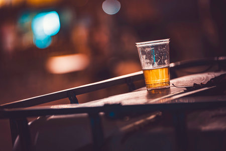 Half-filled transparent plastic cup with beer standing on a wooden table at night, featuring a blue glowing bokeh in the background, symbolizing a party conclusion or social gatheringの写真素材