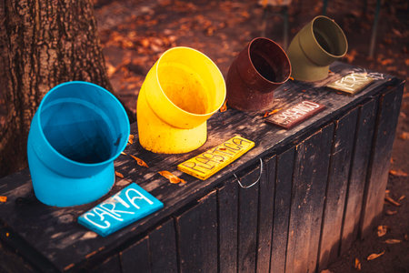 Colorful recycling bins on a wooden stand in a leafy park during autumn, clearly labeled for paper,...