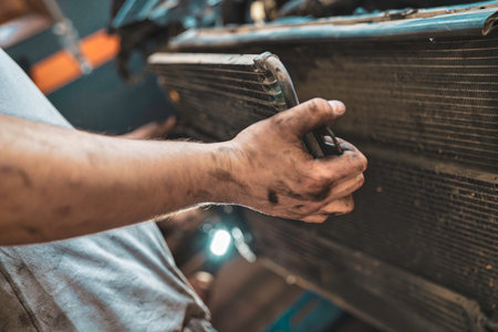 Mechanic with grimy hands holds a car radiator while inspecting the engine during routine maintenance and repair in a busy garage workshop, focused and skilledの写真素材