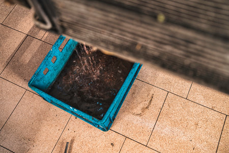 Water pours into a discarded blue plastic crate positioned on light brown tiled flooring, with a blurred wooden structure partially obscuring the top of the frameの写真素材
