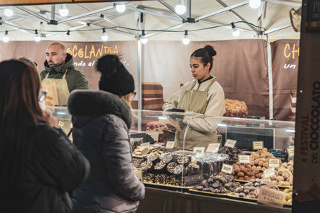 Rovigo, Italy 18 December 2025: people browsing and buying delicious artisan chocolate truffles and sweets from a vendor stall at an outdoor night marketのeditorial素材