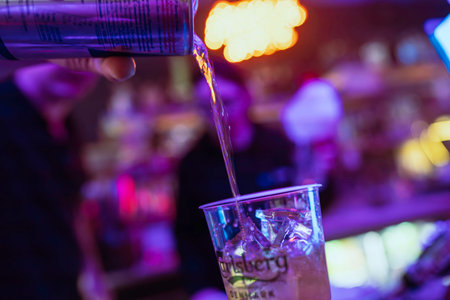 Rovigo, Italy 18 December 2025: bartender pouring chilled drink from bottle into ice-filled plastic cup under purple bokeh lights at lively nighttime bar partyのeditorial素材